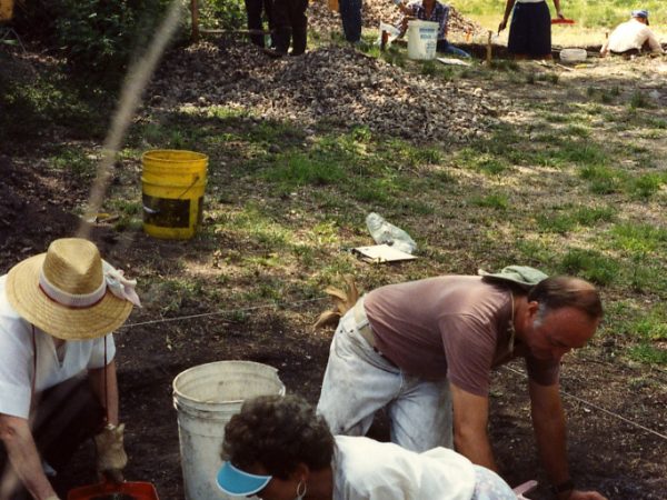 three people in a Archaeological pit, other pit and more people can be seen in the background
