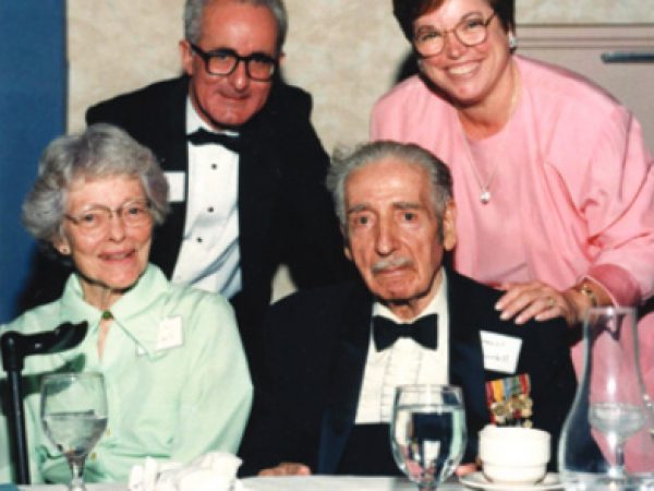 Four people, two seated at a table and two behind them, at a black-tie banquet. All are looking at the camera