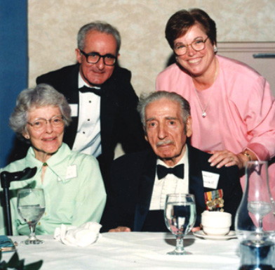 Four people, two seated at a table and two behind them, at a black-tie banquet. All are looking at the camera
