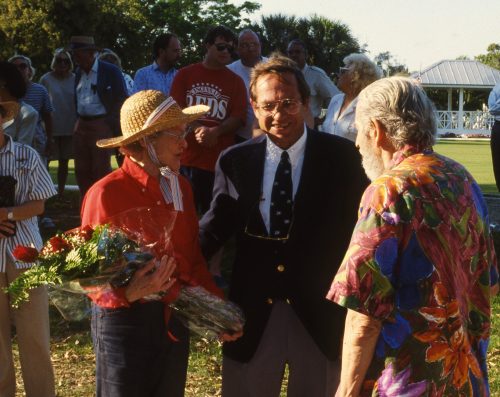 woman in a straw hat holds a bouquet of flowers, she stands next to a man in a suit and another man in a brightly colored shirt