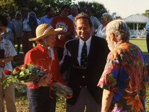 woman in a straw hat holds a bouquet of flowers, she stands next to a man in a suit and another man in a brightly colored shirt