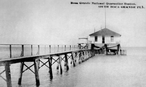 Black and white photo of a house on stilts. A long pier leads to the house