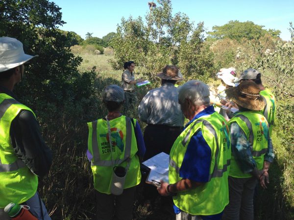 people wearing heritage scout vests stand in tall grass and listen to woman with clipboard