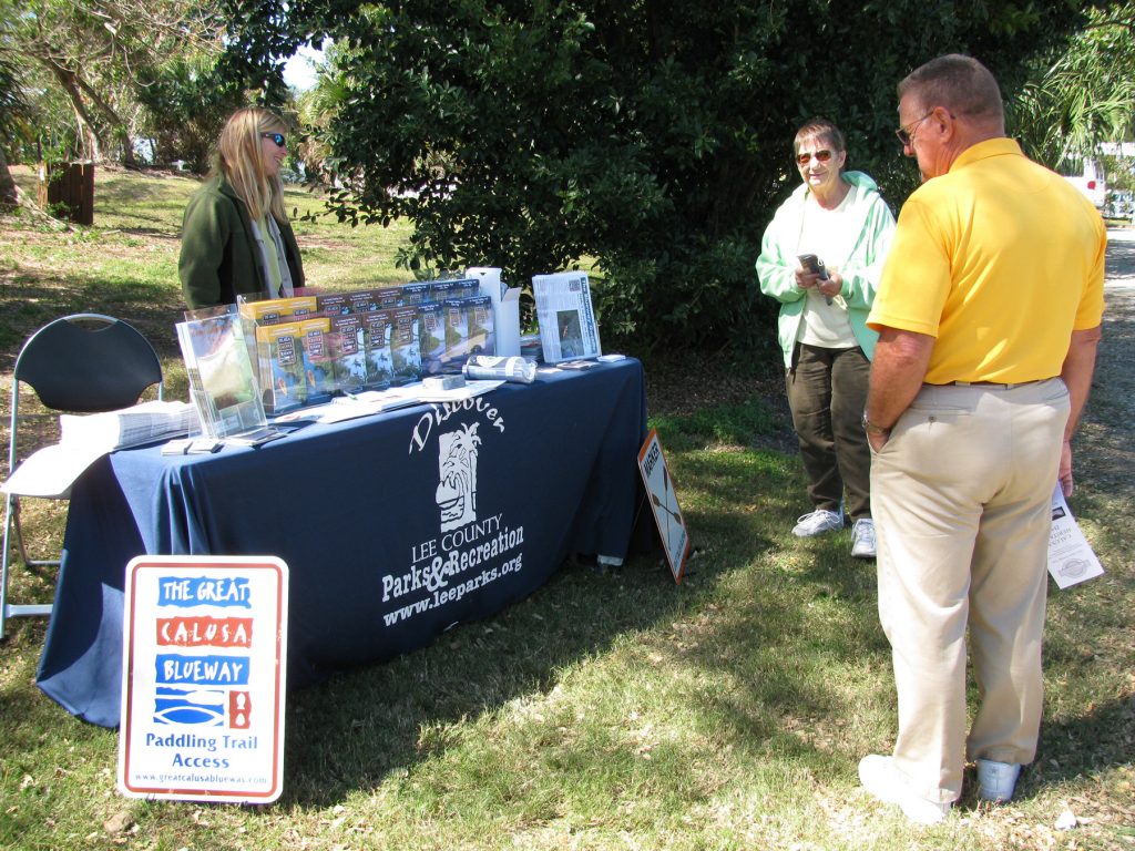person stand behind a display table for Lee County Parks & Recreation and speaks with two people standing infant of the table
