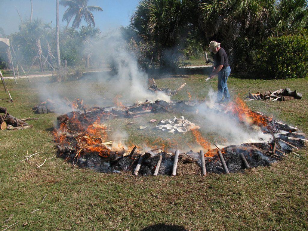 man stands next to a wide ring of fire, at the center of the ring is a pile of pottery
