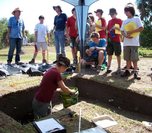 Researcher stands in excavation site while boy scouts stand around the side watching.