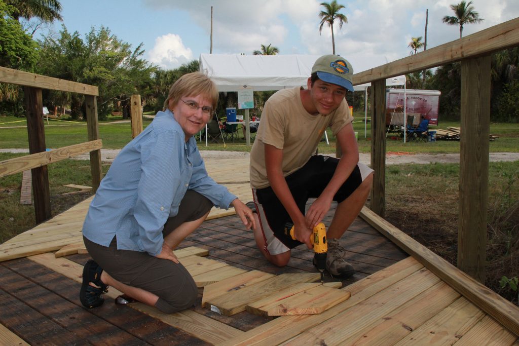 Two people kneel on a footbridge, one holds a electric drill and boards are placed on the footbridge to repair it