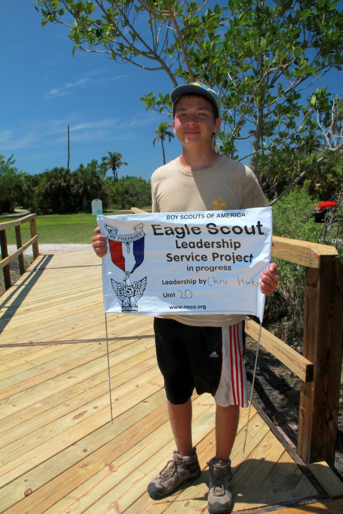 boy stands on the finished footbridge and holds banner that says Eagle Scout leadership service in progress