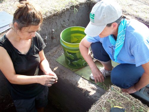 Researcher stands in excavation hold and speaks with another researcher who is kneeling at the edge of the hole.