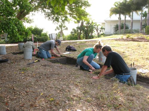 three people sit in shallow, neatly dug holds working on the excavation 