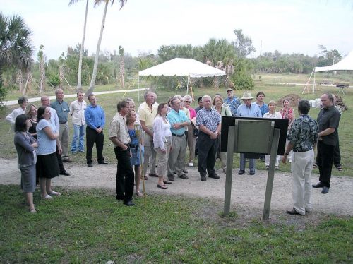 Many people stand before a wooden sign and listen to a person speaking