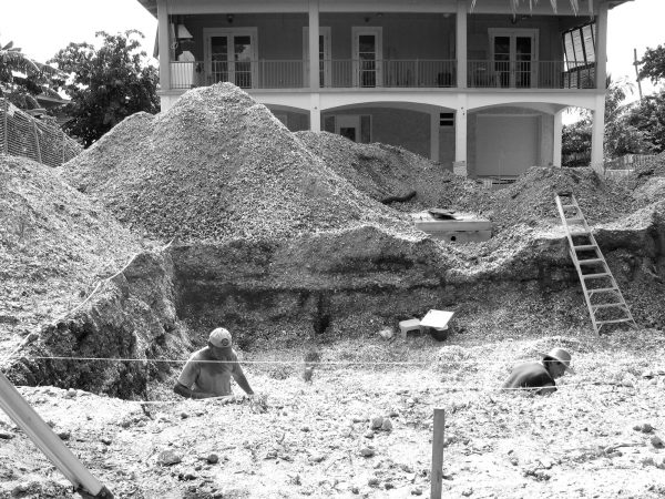 Serval people dig through a large pit and mounds of dirt piled outside a two-story house. One of the mounds of dirt reached the top of the first story. A ladder leans against the side of the pit.