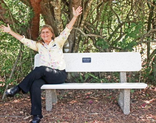 Cindy Bear sitting on a bench smiling with her arms raised. A plaque in the bench reads “Cindy Bear with Love and Respect from your RRC Family.