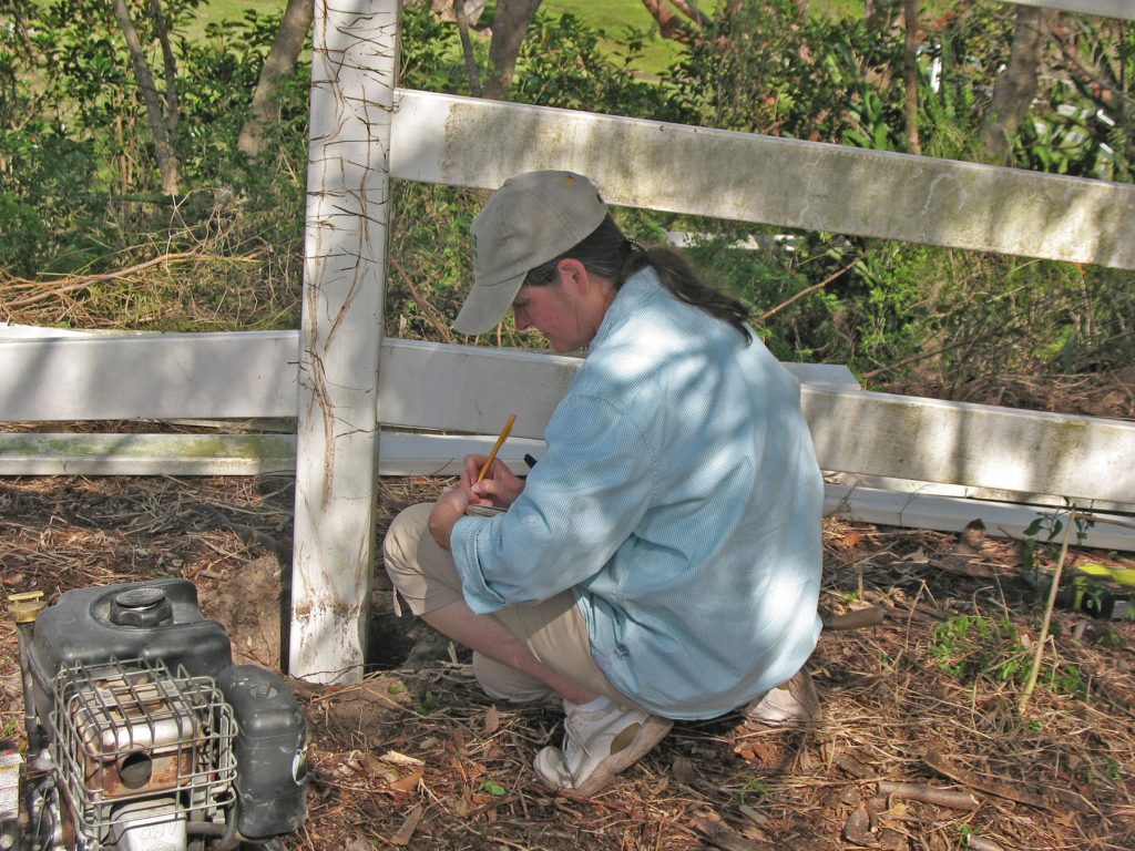 woman kneels next to a fence post and writes in a note book