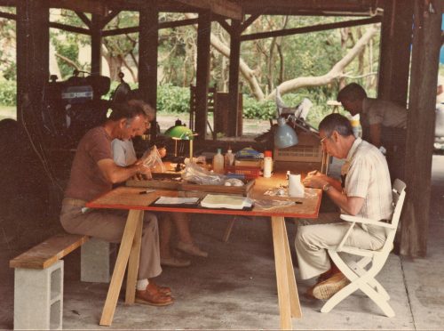 people sit around a sawhorse/plywood table sorting through artifacts.