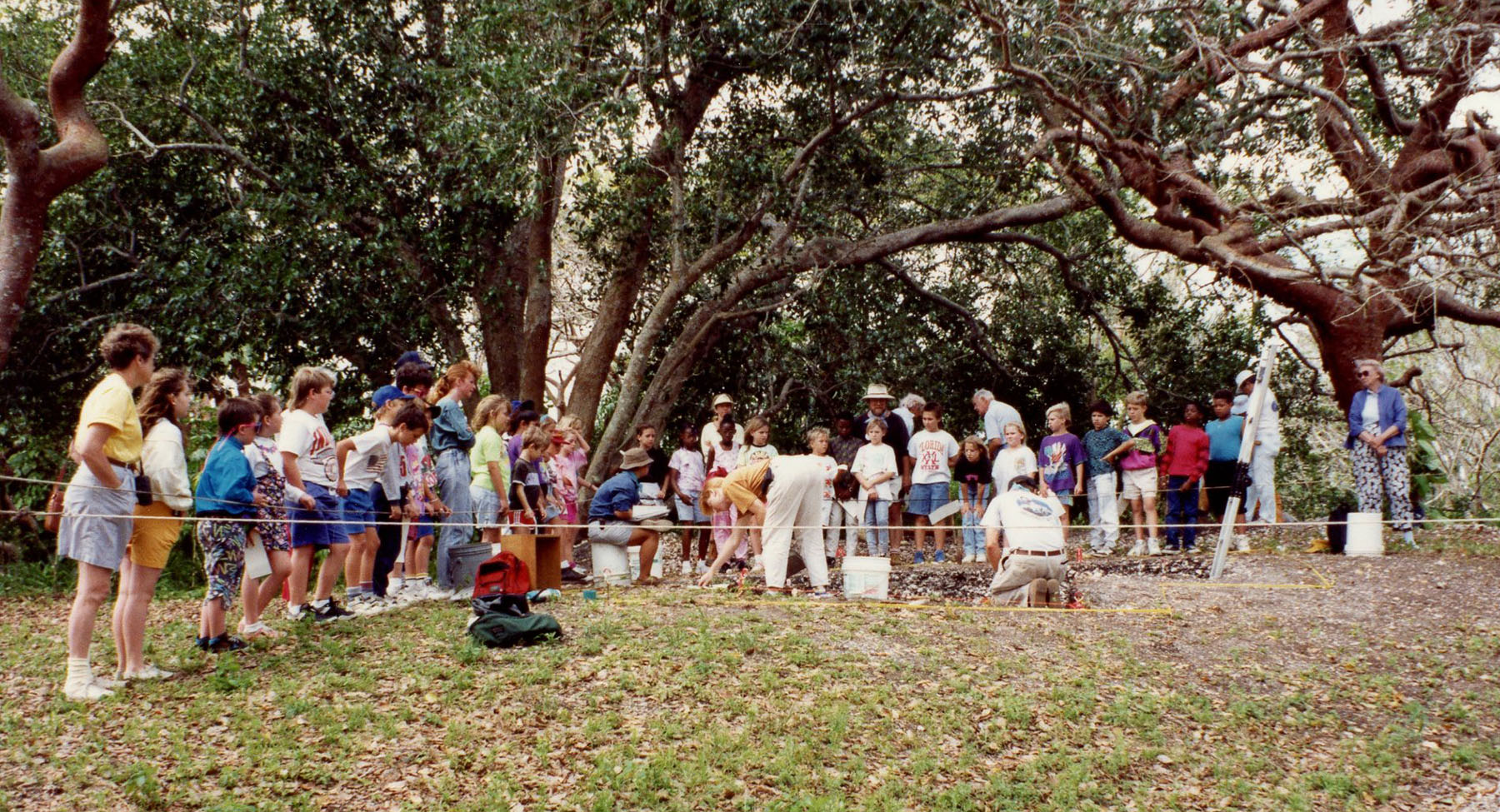 a large group of school children stand around an excavation site and learn about the site.
