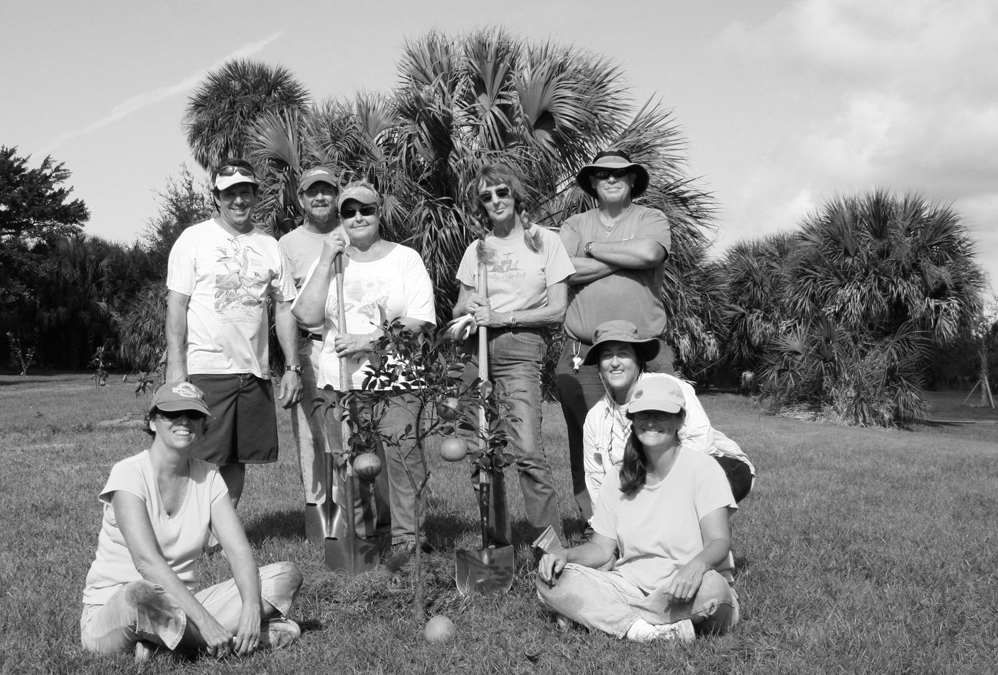 group of people eight stand and sit around a small citrus tree that has just been planted