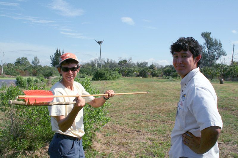 person holding a long throwing stick with feathers on one end. The stand next to a person in an RRC shirt.