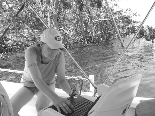 woman sitting on a boat working on a laptop mangrove trees and water can be seen behind her