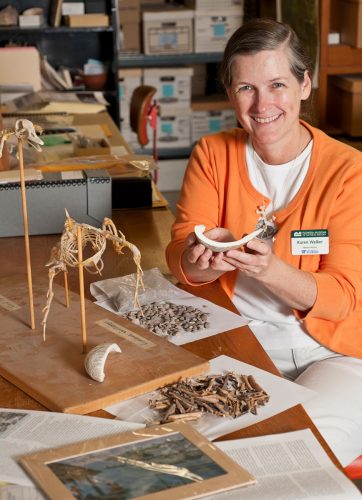 woman sits at a desk spread with many specimens. she is holding a shell and smiling at the camera