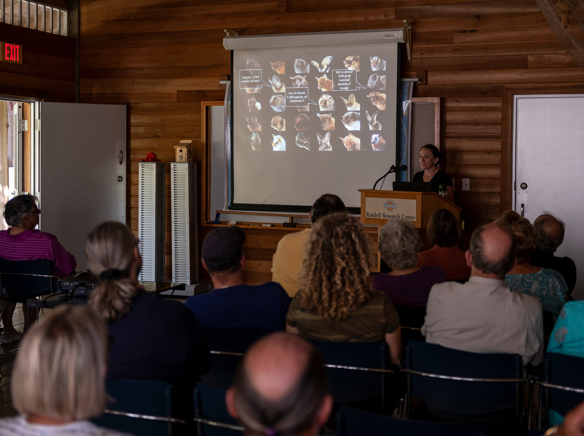 researcher stands behind a podium with a Randell Research Center and speaks to an audience. A projection screen behind her shows many photos of bats.