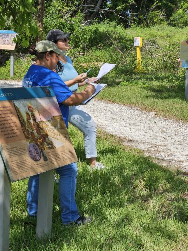 two people stand in the grass next to a gravel path, both hold note books and pens