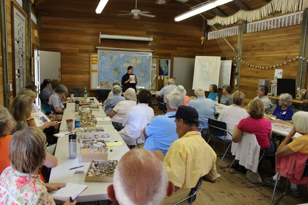 a person stands in front of a map and speaks to a large group of people seated at two long white tables