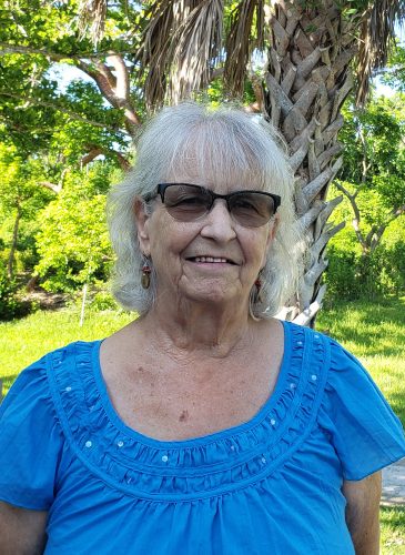 potrait of an older woman in a blue shirt, behind her are green trees and grass