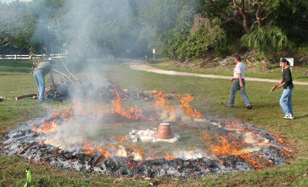 several people stands next to a wide ring of fire, at the center of the ring is a pile of pottery