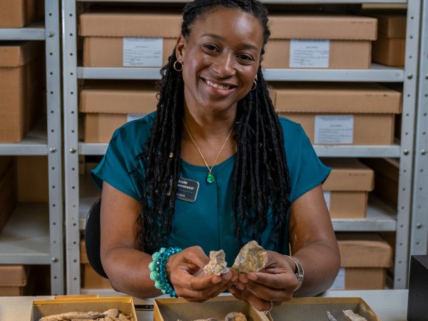 Nicole Fuller smiling at the camera holding specimens and sitting behind a table covered with boxes filled with more museum specimens