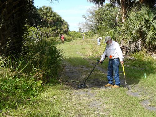 people waling along dirt and grass covered road holding metal detectors 