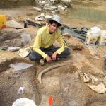 Student with yellow shirt sitting in a fossil site with an elephant relative rib in from of him