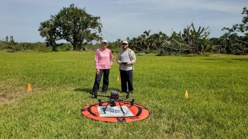 two people stand behind a drone seated on a portable circular landing pad one person is holding the drone remote controls