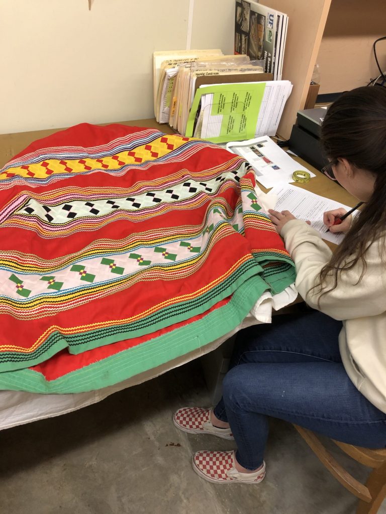 a person is sitting at a desk to write notes on paper with a colorful garment spread out in front of her primarily in red with detailed bands of colors and quilted patterns in geometric shapes with green and white and yellow and black