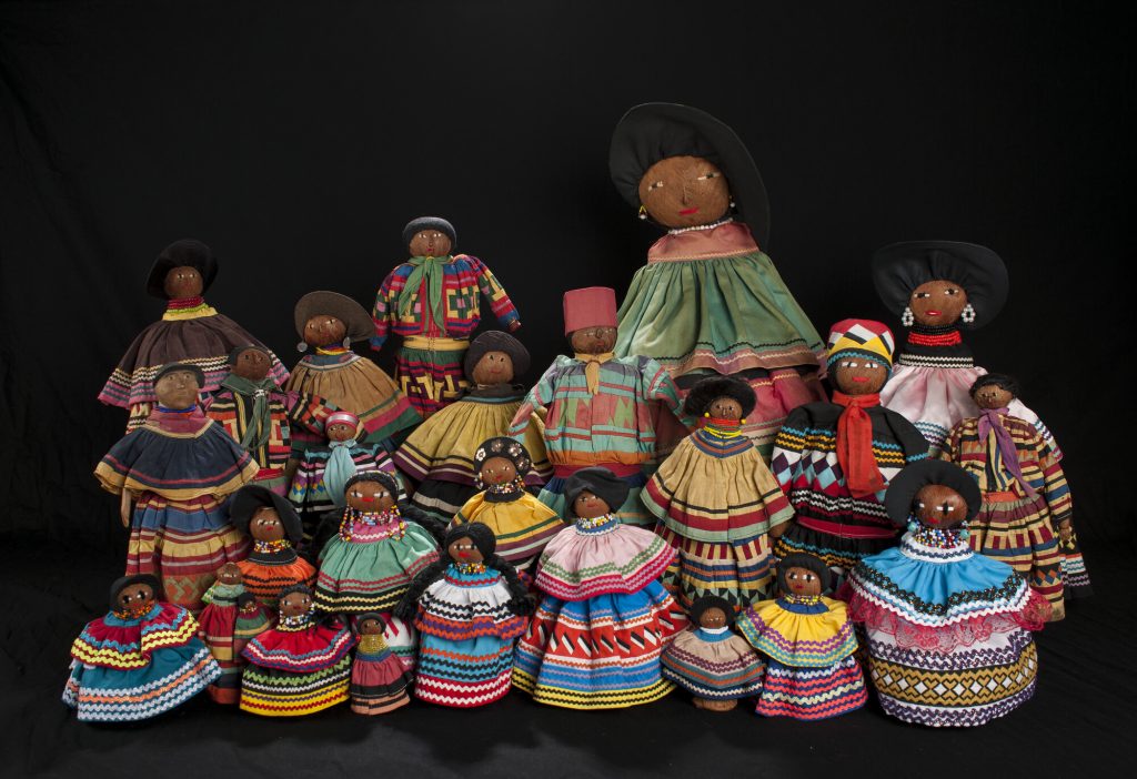 a collection of dolls in several sizes are positioned standing in several rows to show their colorful dresses with bright rickrack and quilted stripes and each hand-made face on brown fibrous material