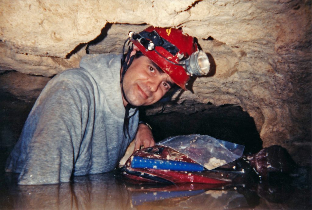 man in a cave wearing a red hard hat with light and standing in water up to his chest
