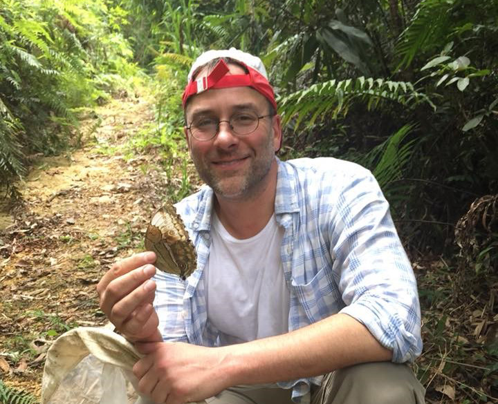 a person is crouching in a forest and holding up a large butterfly towards the camera while smiling
