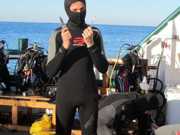 françois standing on the boat deck in his wetsuit with his hood pulled up over his nose and mouth and holding a dive knife