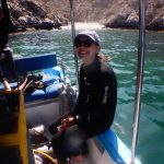 woman in wetsuit and cap smiling on a boat with water and rocks and a little sandy beach in the background