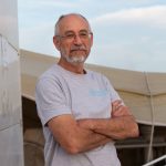 man in grey tshirt with arms crossed and looking at the camera on the deck of a large boat