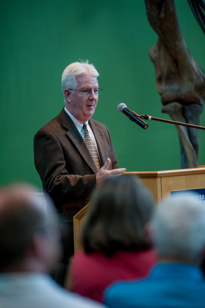 man in brown suit stands behind a podium and microphone and speaks to an audience