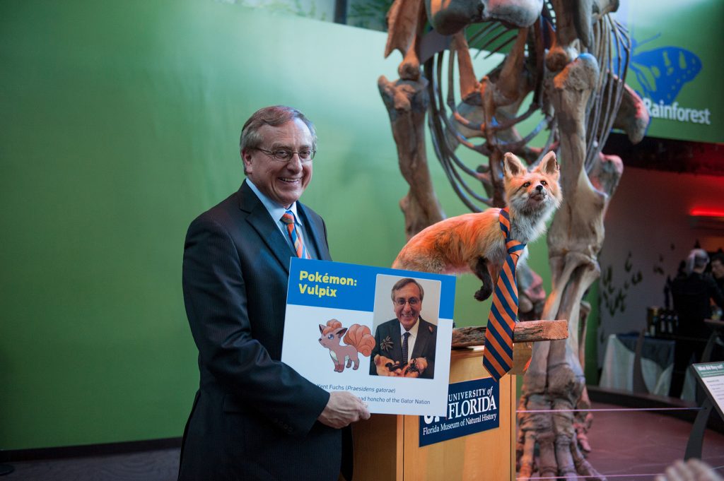 Kent Fuchs stand in front of the Florida Museum Mammoth and a fox specimen that is wearing a orange and blue tie.