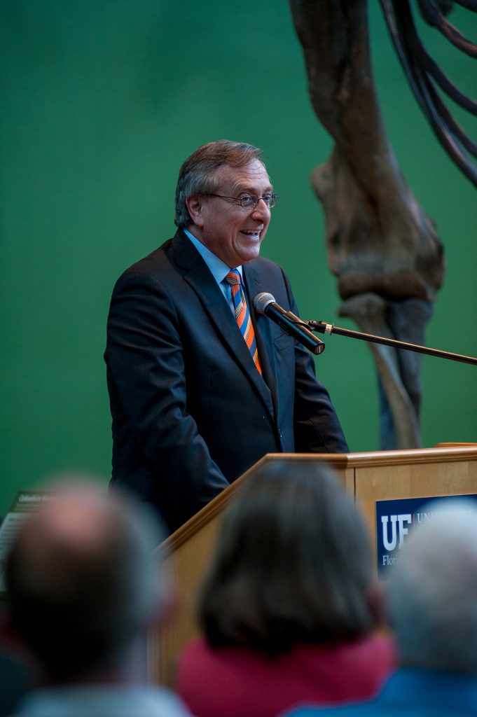President Fuchs in blue suit and orange and blue tie stands behind a podium and microphone and speaks to an audience
