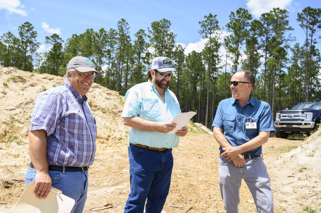 three men stand at the edge of a fossil dig site surrounded by pine trees