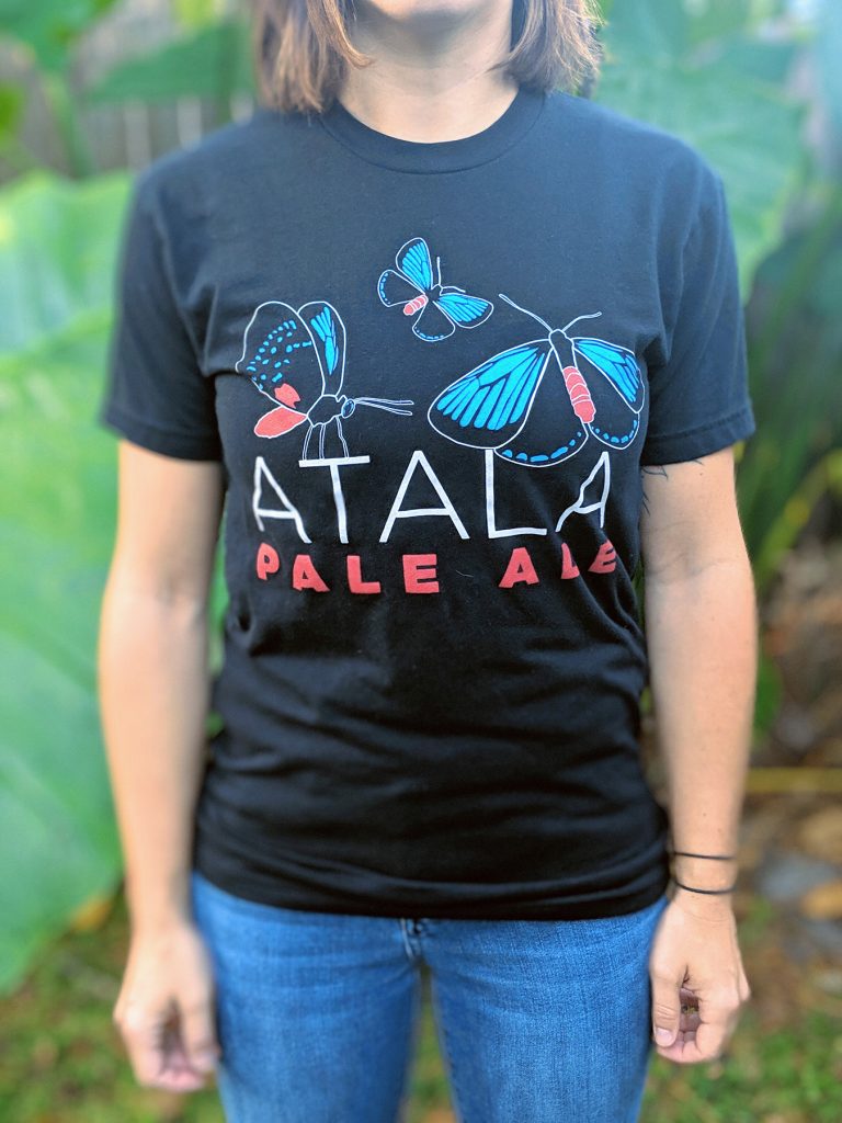 Person wearing a Atala Pale Ale T-Shirt. Black t-shirt with black, blue and red butterflies and "Atala Pale Ale" in white and red