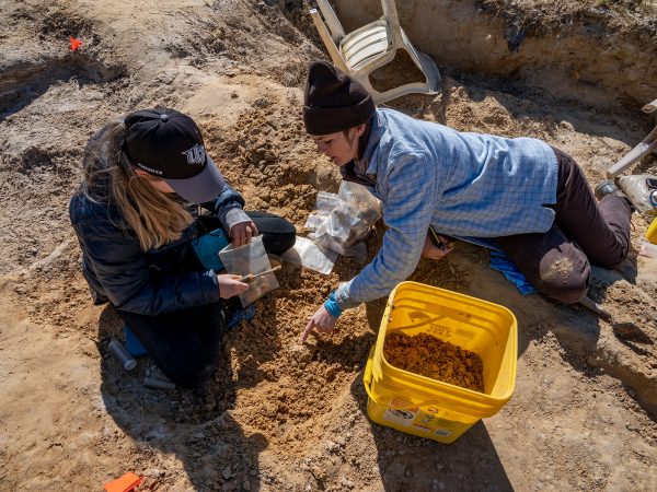 Photographed from above, one teacher kneels on the ground and points to something recently uncovered in the red dirt. Another teacher holds a screwdriver and plastic collection bag