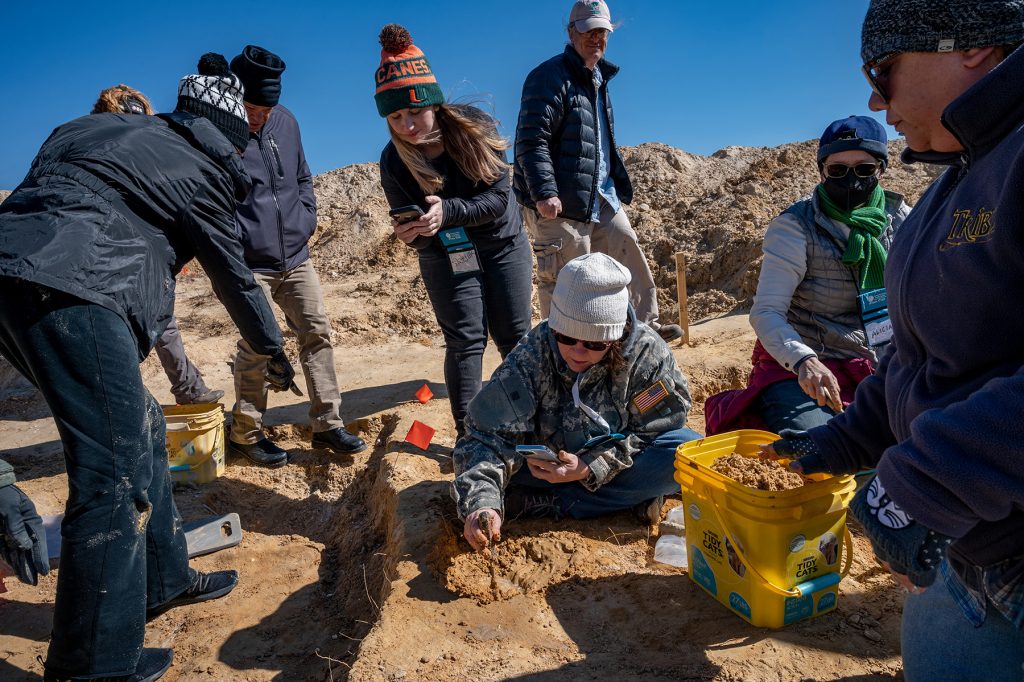 person in the center of the photo sits on the ground next to the fossil she has just uncovered, she holds a dirt covered screwdriver to point at the fossil and a phone that she is using to take a picture. Around her other participants lean in taking their own photos