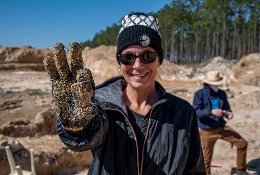 Visiting teacher holds up a two inch fossil close to the camera. The fossil and her glove are covered in dirt. She is smiling at the camera
