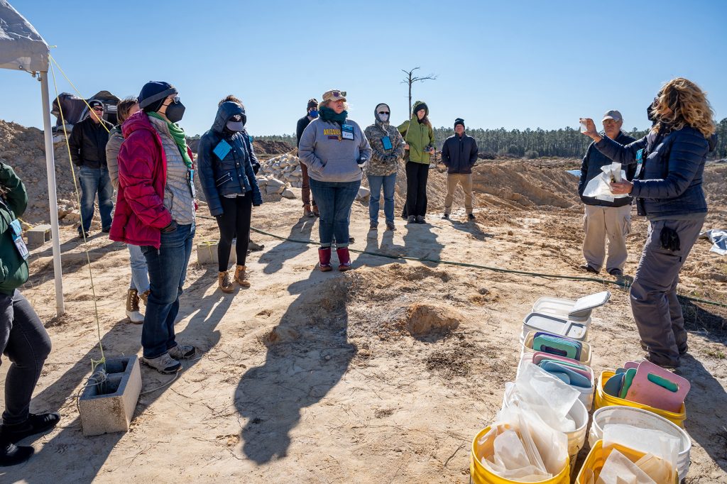 Many people sanding in an open dirt covered field. One researcher on the right holds up a plastic container and bags explaining the tools used in the fossil dig to the other people listening. Everyone is wearing heavy coats, hats, and masks