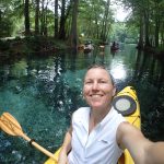 a person takes a selfie from a kayak with a freshwater spring behind her surrounded by trees